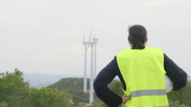 Üniformalı ve kasklı kadın elektrik mühendisinin dağlardaki rüzgar türbinlerine bakışı.
