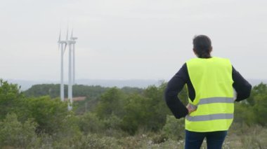 Üniformalı ve kasklı kadın elektrik mühendisinin dağlardaki rüzgar türbinlerine bakışı.