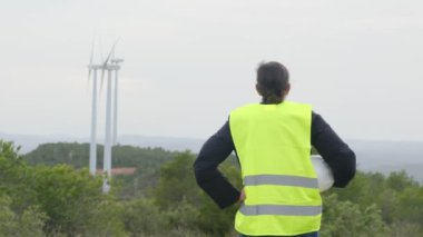 Üniformalı ve kasklı kadın elektrik mühendisinin dağlardaki rüzgar türbinlerine bakışı.
