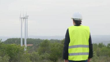 Üniformalı ve kasklı kadın elektrik mühendisinin dağlardaki rüzgar türbinlerine bakışı.