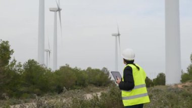 Kadın elektrik mühendisi telsizle konuşuyor ve dağlardaki rüzgar türbinlerinin yanında dijital tablet kullanıyor.