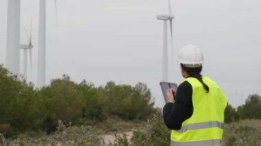 Kadın elektrik mühendisi telsizle konuşuyor ve dağlardaki rüzgar türbinlerinin yanında dijital tablet kullanıyor.