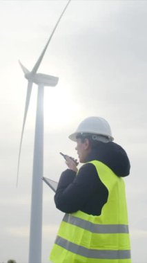 Kadın elektrik mühendisi telsizle konuşuyor, dijital tablet kullanıyor ve rüzgar türbinini kontrol ediyor.