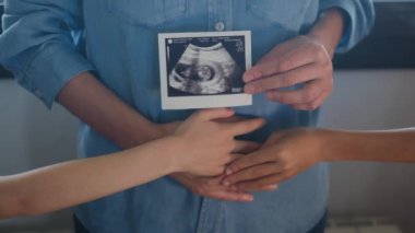 Kısmen müstakbel bebeğinin ultra sesli resmini gösteren ve hamile karnına çocuklarıyla dokunan kadının görüntüsü. 