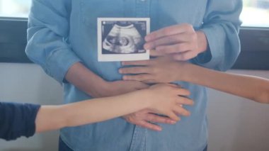 Kısmen müstakbel bebeğinin ultra sesli resmini gösteren ve hamile karnına çocuklarıyla dokunan kadının görüntüsü. 