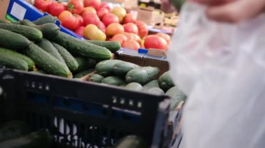 Elinde çanta olan bir kadın ve markete salatalık koyuyor.