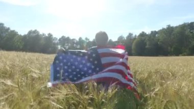 Buğday tarlasında koşan Amerikan bayrağı taşıyan çocuğun arka görüntüsü. 4 Temmuz Bağımsızlık Günü.