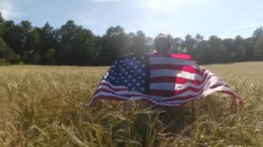 Buğday tarlasında yürüyen Amerikan bayrağı taşıyan bir çocuğun arka görüntüsü. 4 Temmuz Bağımsızlık Günü.