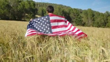 Buğday tarlasında yürüyen Amerikan bayrağı taşıyan bir çocuğun arka görüntüsü. 4 Temmuz Bağımsızlık Günü.