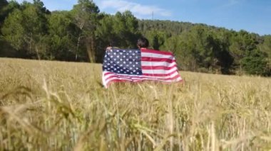 Buğday tarlasında yürüyen Amerikan bayrağı taşıyan bir çocuğun arka görüntüsü. 4 Temmuz Bağımsızlık Günü.