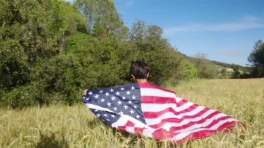 Buğday tarlasında yürüyen Amerikan bayrağı taşıyan bir çocuğun arka görüntüsü. 4 Temmuz Bağımsızlık Günü.