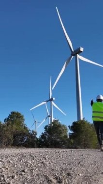 Dijital tabletli elektrik mühendisleri üniforma giyip, güneşli bir günde rüzgar türbinlerini kontrol ediyorlar.