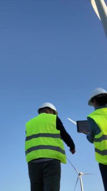 Dijital tabletli elektrik mühendisleri üniforma giyip, güneşli bir günde rüzgar türbinlerini kontrol ediyorlar.