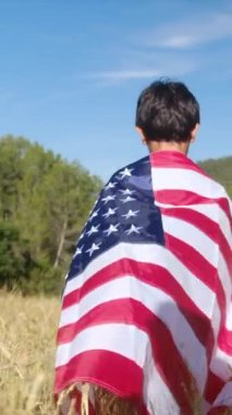 Buğday tarlasında yürüyen Amerikan bayrağı taşıyan bir çocuğun arka görüntüsü. 4 Temmuz Bağımsızlık Günü.