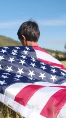 Buğday tarlasında yürüyen Amerikan bayrağı taşıyan bir çocuğun arka görüntüsü. 4 Temmuz Bağımsızlık Günü.