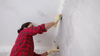 Kırmızı gömlekli yüz maskesi takan ve yenileme dairesinde duvarları yıkayan bir kadın. Yavaş çekim videosu.