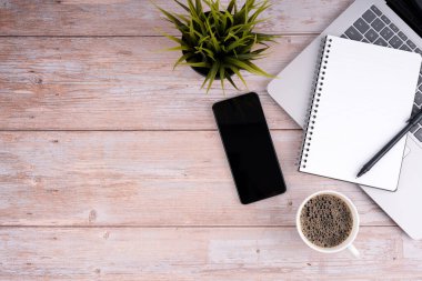 Bir dizüstü bilgisayar, bir defter, bir kalem, bir cep telefonu, bir fincan kahve ve ahşap bir masanın üzerinde saksı yeşil bir bitki.