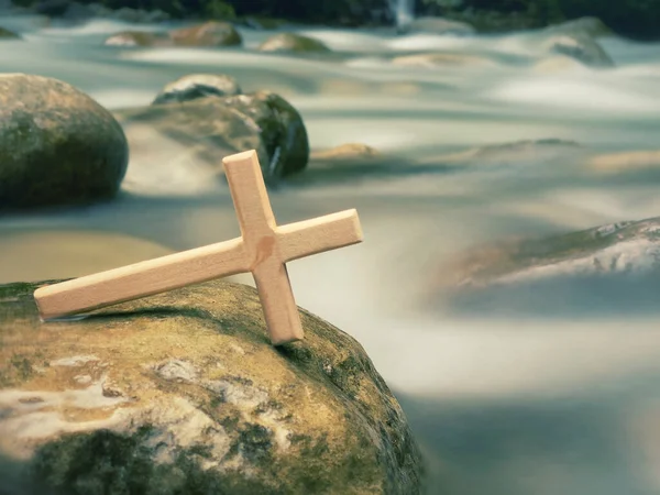 Wooden cross leaning on rock with river background. Christianity, easter, baptism concept.