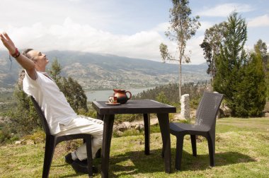 Beyazlar içinde bir adam dağın tepesinde bir sandalyeye oturmuş sıcak bir kahve içiyor ve güneşin doğuşunu seyrediyor.