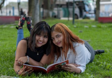 Parkta çimlerin üzerinde yatan iki arkadaş gülümseyip kitap okuyorlar. Yüksek kalite fotoğraf
