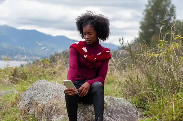 afro girl checking her cell phone before a live broadcast sitting on a mountain rock. High quality photo