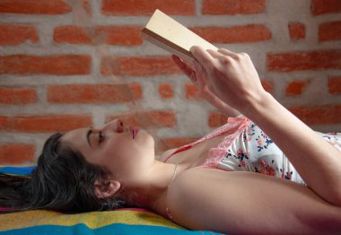 attractive young woman from ecuador lying on her back on her bed in her nightclothes reading a book attentively in the morning after waking up. book day. High quality photo clipart