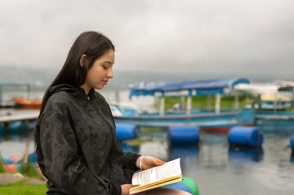 attractive young man from latin america sitting by a pier with boats reading a book while it rains. book day. High quality photo