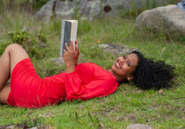 Genç Latin Amerikalı kız sırtüstü uzanıp elinde açık bir kitapla güzel bir gülümsemeyle kameraya bakıyor. Kitap günü. Yüksek kalite fotoğraf