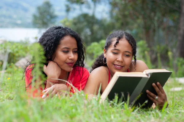 two latin sisters reading a book in a peaceful and quiet environment surrounded by trees and nature near a lake. book day. High quality photo
