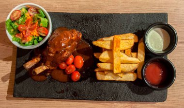 Patates kızartması, salata, kiraz domatesleri, ketçap ve mayonez ile servis edilen lezzetli osso buco tabağı ahşap bir masada yenmeye hazır.