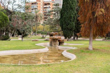Güneşli bir günde çevredeki binaları yansıtan modern bir fıskiyede akan su yeşil ağaçlar ve çimenlerle huzurlu bir atmosfer yaratır.