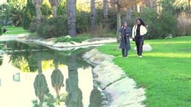 Paltolu iki iş kadını, bereketli yeşil bir parktaki dingin bir göletin yanında yürürken, doğanın güzellikleriyle dolu güneşli bir günün tadını çıkarıyorlar.