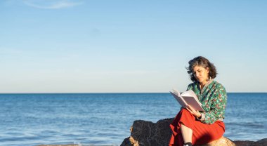 Güneşli bir günde, sakin bir denizde, olgun bir kadın deniz kenarındaki bir kayanın üzerinde oturup, huzurlu ve rahatlatıcı bir sahne yaratarak kitap okumaktan zevk alır.