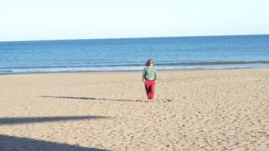 Kumlu sahil şeridinde sakin bir şekilde yürüyen, sıcak güneş ışığında güneşlenen, canlı kırmızı pantolon ve yeşil gömlek giyen, huzurlu deniz kenarı tatil havasını somutlaştıran bir kadın.