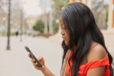 Canlı kırmızı bir elbise giyen genç Afrikalı bir kadın, güneşli bir bahar gününde şehir merkezinde akıllı telefonunu sosyal medyada gezinirken bir bankta oturuyor.