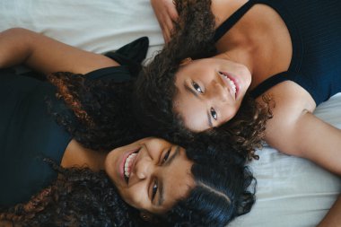 İki neşeli kıvırcık saçlı genç kız, bir yatağa uzanmış, kolsuz tişört giyiyor, gülümsüyor ve kahkahalarını paylaşıyor, birlikte kaygısız anların tadını çıkarıyorlar, yukarıdan yakalanmışlar.