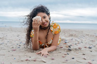Sıcak kumda uzanan turist, gözünün dibinde bir deniz kabuğu tutuyor ve güneşli bir yaz tatili boyunca kumsalda neşeyle gülümsüyor, doğanın güzelliğini ve rahatlığını kucaklıyor.