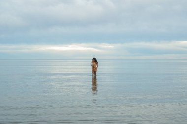 Sakin denizde elinde antika bir kamerayla yürüyen genç bir kadın bulutlu bir günde tatilin tadını çıkarıyor sakin bir su ve yüzeyde yansıyan ışıkla.