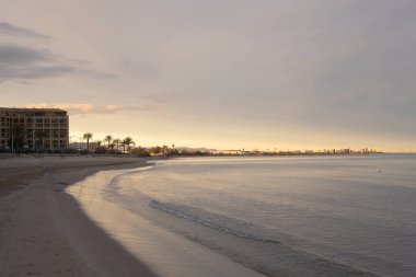 Gün doğumunda bir tatil köyünün önündeki kumlu sahilde hafif dalgalar yavaşça süzülüyor. Arka planda muhteşem Cullera şehri, Valencia, İspanya.