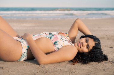 Genç turist kumlu sahilde uzanıp yaz tatilinde güneşin tadını çıkarıyor. Canlı bir çiçekli mayo giyip, elini başının arkasında dinlendiriyor.