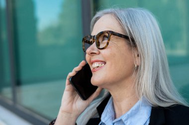 Gri saçlı ve gözlük takan mutlu bir iş kadını cep telefonuyla konuşuyor. Çağdaş cam binanın yanında duruyor ve hoş sohbetler ediyor.