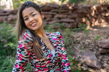 Neşeli genç kadın renkli bir elbise giyerken gülümsüyor, sakin bir ormanda zarifçe poz veriyor, arkasında büyüleyici bir taş duvarla güneşli bir günün tadını çıkarıyor.