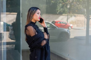 Modern bir ofis binasının önünde duran genç bir iş kadını şık siyah giysiler içinde, akıllı telefonuyla konuşuyor.