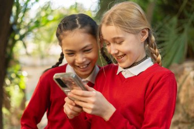 Okul üniforması giyen iki genç kız öğrenci okul bahçesinde tatillerinin tadını çıkarırken akıllı telefondan eğlenceli videolar veya fotoğraflar izliyorlar.