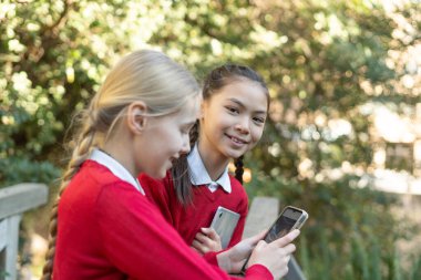 Okul üniformalı iki genç öğrenci açık havada akıllı telefonları kullanarak, sosyal medyayla iletişim kurarken gülümsemelerini paylaşıyorlar ve canlı bir okul bahçesindeki son teknolojinin tadını çıkarıyorlar.