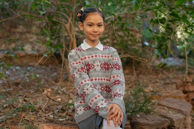 Neşeli genç bir öğrencinin portresi Noel desenli bir süveter ve okul üniforması giyiyor, güneşli bir günde ağaçların arasında oturuyor.