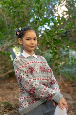 Asyalı bir ilkokul öğrencisi, Noel desenli bir süveter giyer, bir ormanda rahatça oturur, kışın sıcaklığının keyfini çıkarırken hafifçe gülümser.