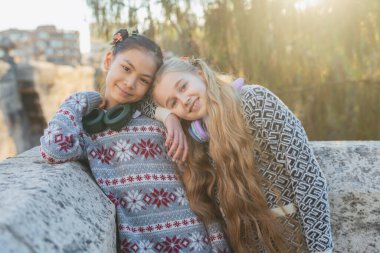 Kış süveteri ve kulaklık takan iki genç kız öğrenci güneşli bir sonbahar gününde parkta birbirlerine yaslanıp gülümsüyorlar.
