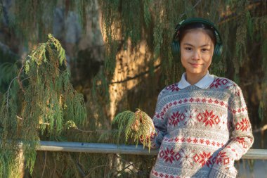 Gülümseyen genç bir öğrenci Noel temalı bir süveter ve kulaklık giyer, güneşli bir parkta yemyeşil ağaçlarla birlikte korkuluklara yaslanırken müziğin tadını çıkarır.