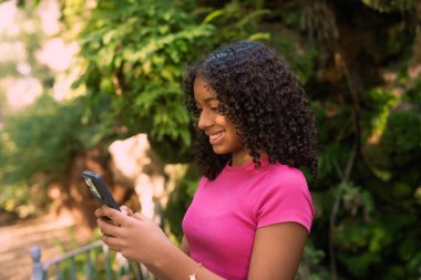 Parkta güneşli bir yaz gününün tadını çıkaran gülümseyen genç bir kadın pembe bir tişört ve gür yeşil ağaçlarla çevrili şık bir bilezik takarken cep telefonunu kullanıyor.